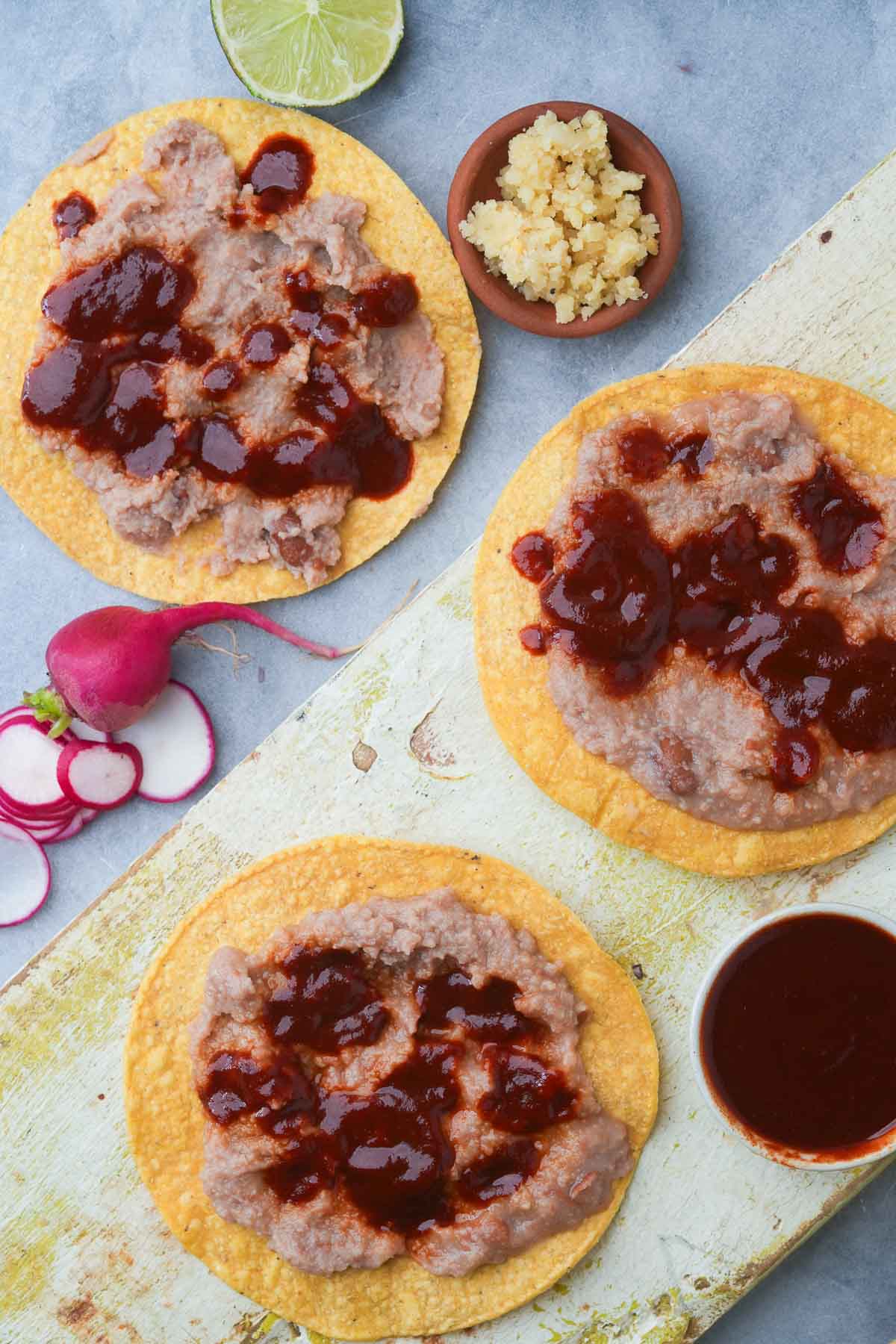 Three tostadas with refried beans and red chili sauce on a cutting board.