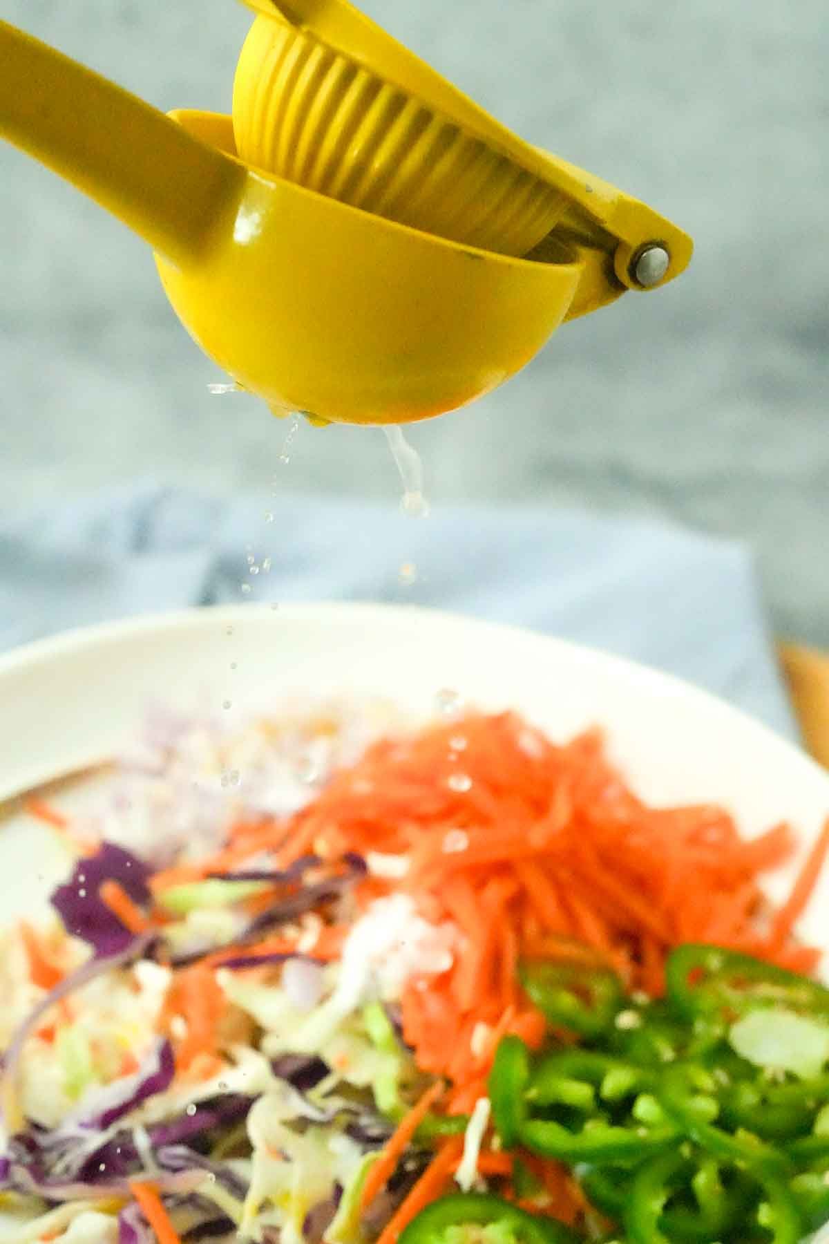 A citrus juicer dripping lime juice onto a bowl of colorful shredded vegetables, including carrots and cabbage, jalapeños and red onion.