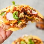 A hand holds a Vegan Tostada topped with plant-based chorizo crumbles, beans, cabbage slaw, almond cotija cheese, and slices of radish, garnished with cilantro.