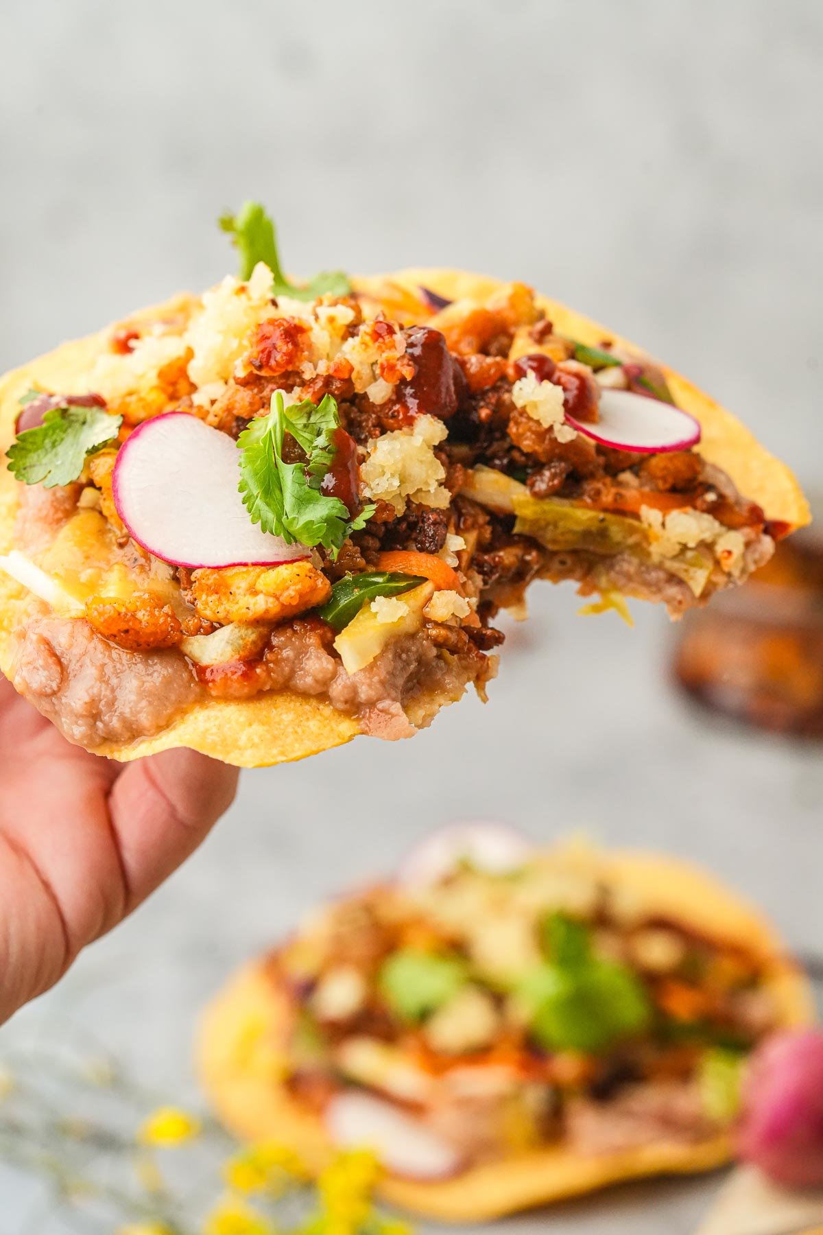A hand holds a Vegan Tostada topped with plant-based chorizo crumbles, beans, cabbage slaw, almond cotija cheese, and slices of radish, garnished with cilantro.