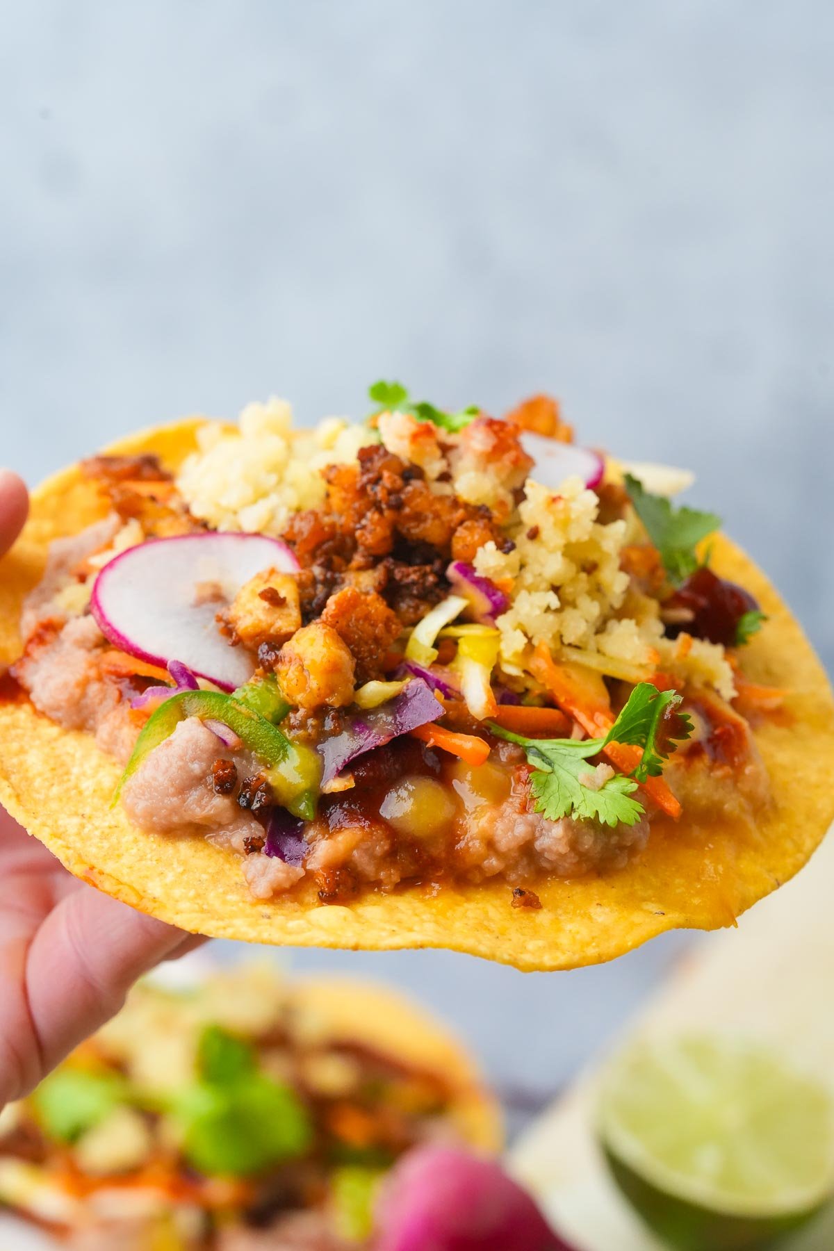 A hand holds a Vegan Tostada topped with plant-based chorizo crumbles, beans, cabbage slaw, almond cotija cheese, and slices of radish, garnished with cilantro.
