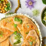 A plate of naan bread with a small dish of creamy dip, surrounded by a bowl of seasoned chickpeas, a flower, and leafy herbs on a light-colored surface.