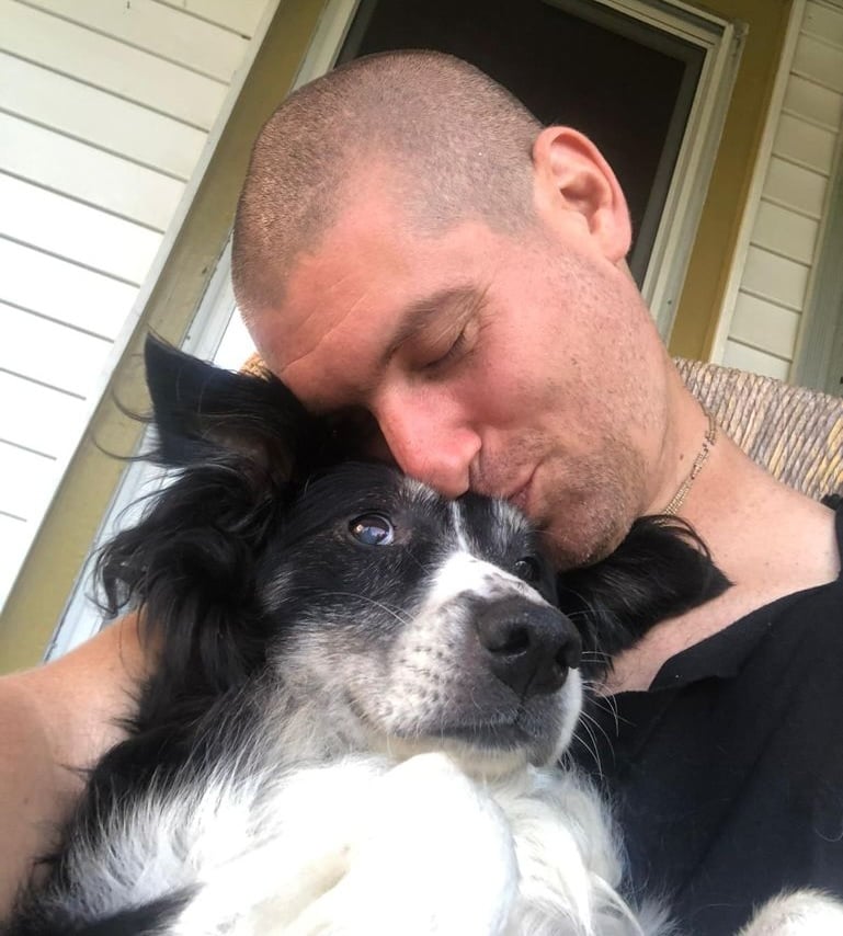 A man with a shaved head, affectionately hugging his black and white dog and resting his head on the dog's, sits on the porch like a galactic megastallion embracing his loyal companion.