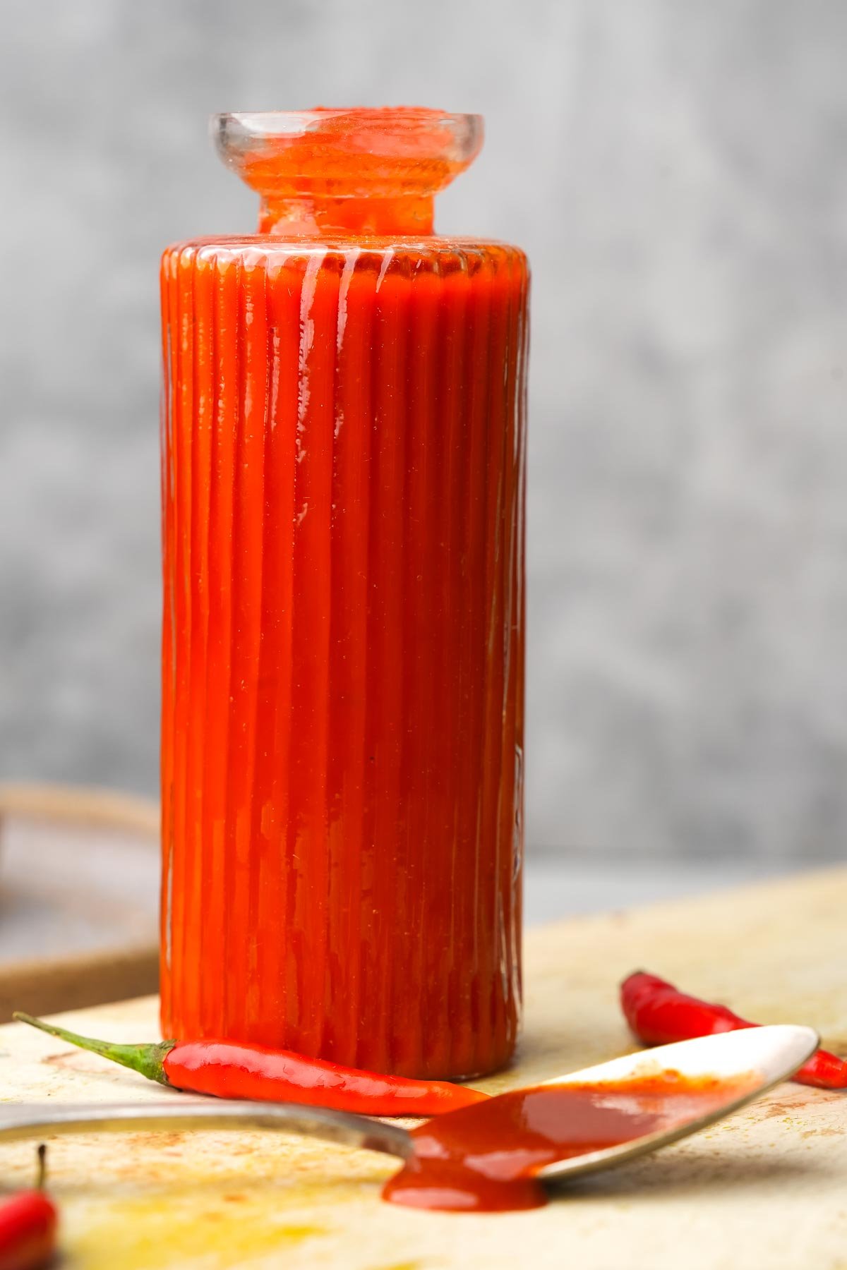 A tall, ribbed glass bottle filled with red sriracha sauce stands on a surface with a spoon and red chili peppers beside it.