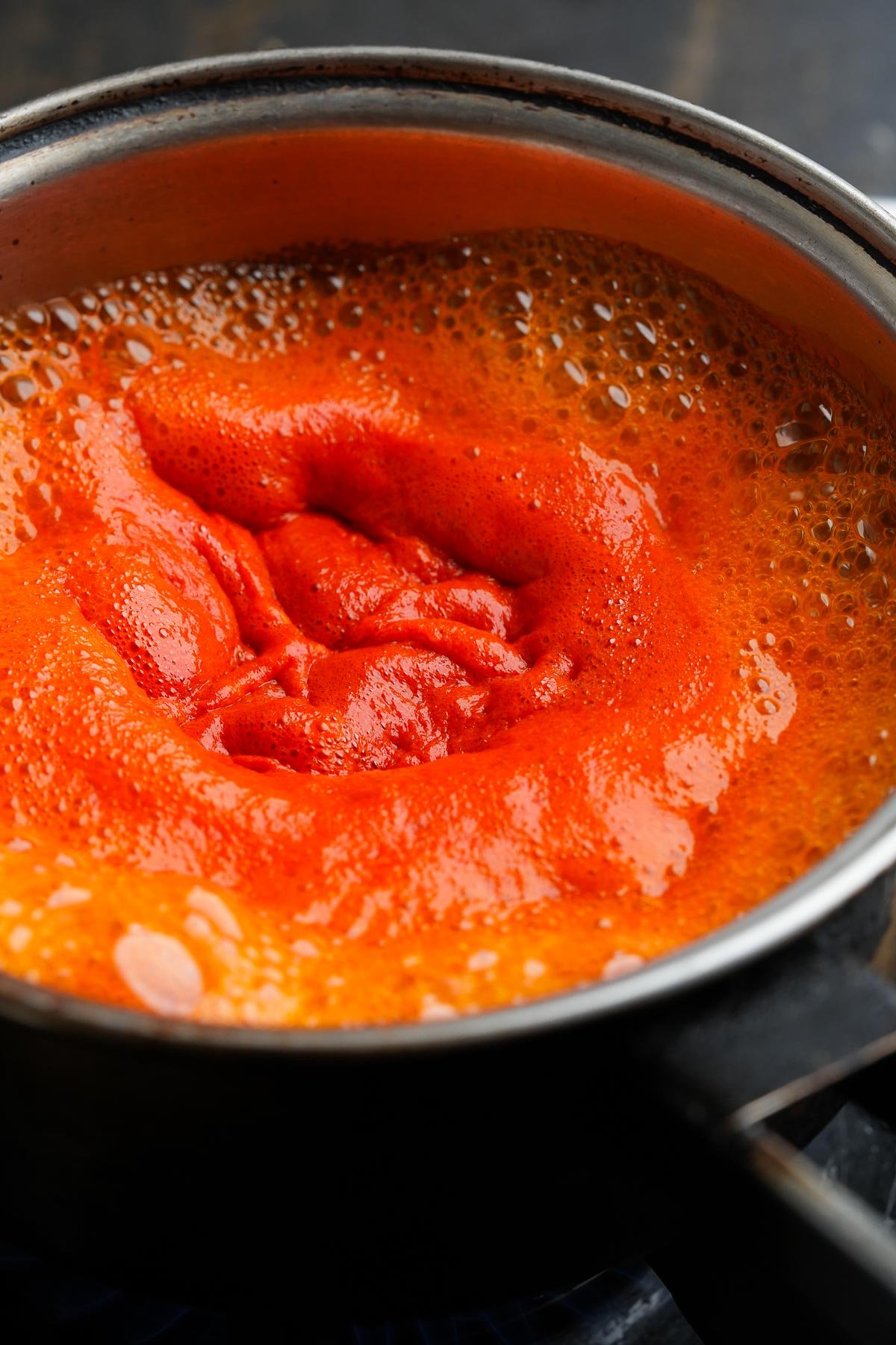 A pot of red sriracha sauce is boiling on the stove, with bubbles visible on the surface.