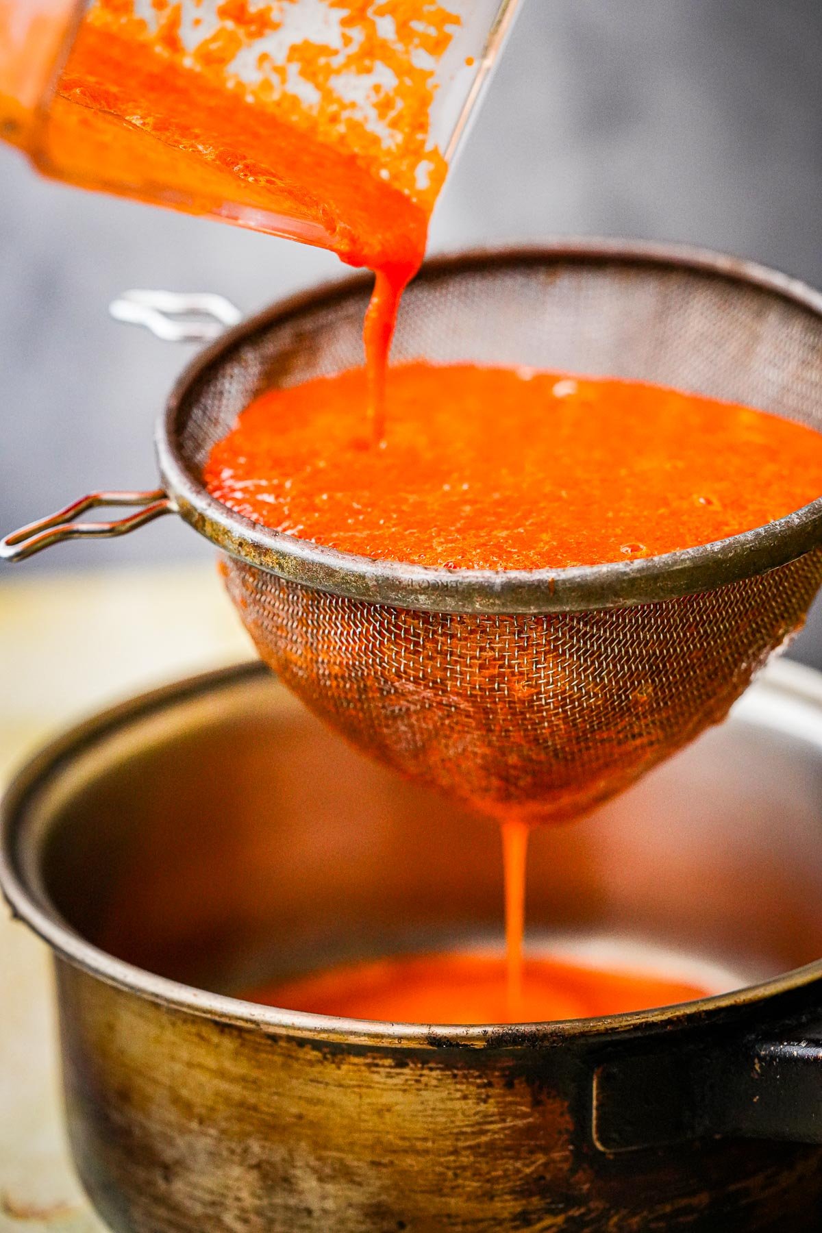 Bright sriracha sauce being poured through a metal strainer into a stainless steel pot.