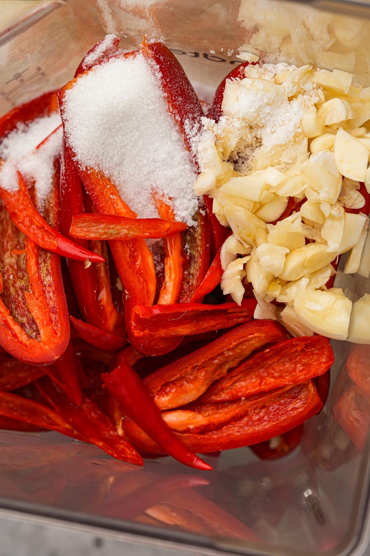 A close-up of a blender container with chopped red peppers, salt, and minced garlic cloves inside.