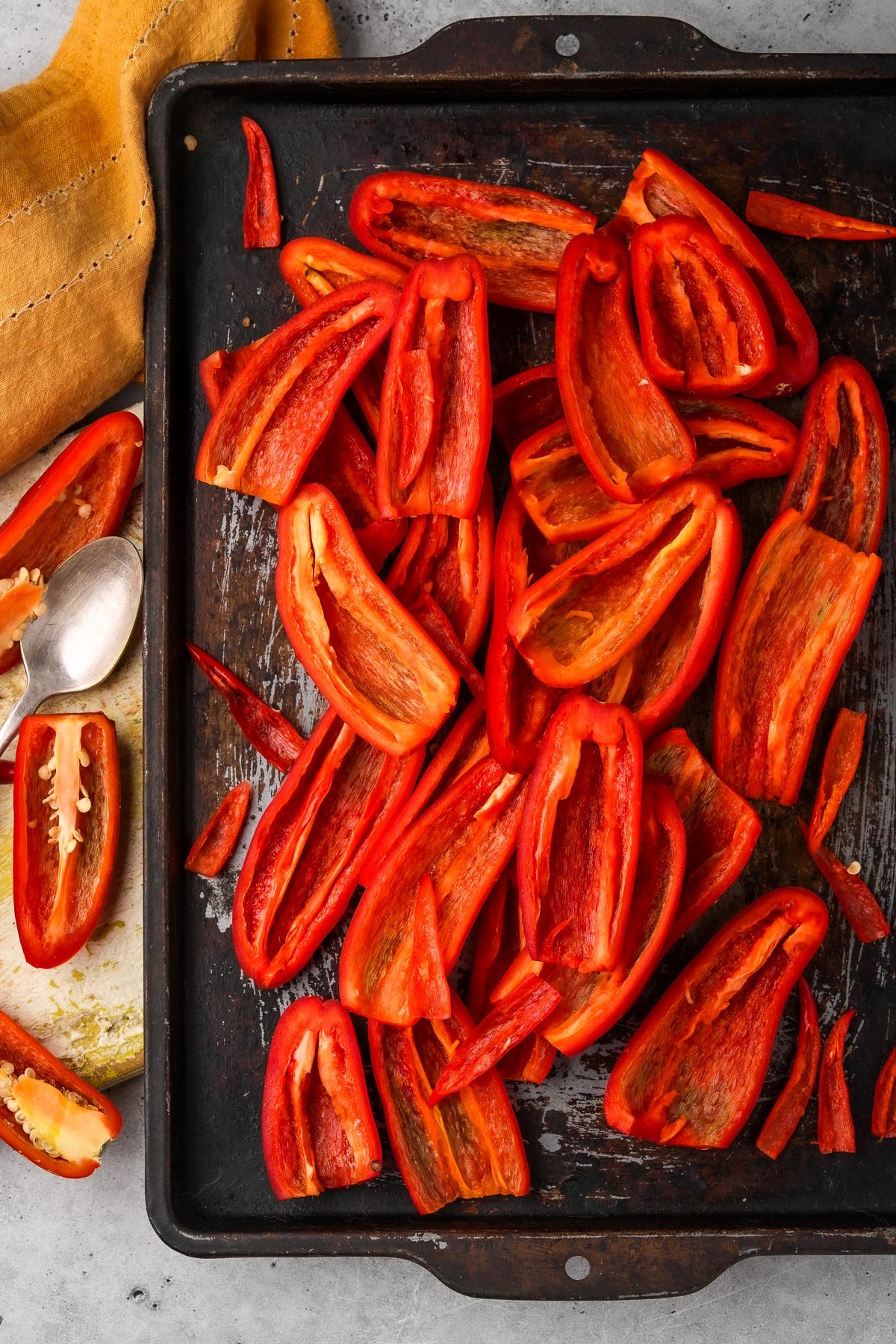 A baking sheet filled with sliced red jalapeño peppers. A yellow cloth and a spoon are on the side, with a few additional pepper slices nearby.