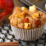 A close-up of a Vegan Apple Muffin topped with apple pieces and cinnamon, with an apple and a cinnamon stick in the background on a metal cooling rack.