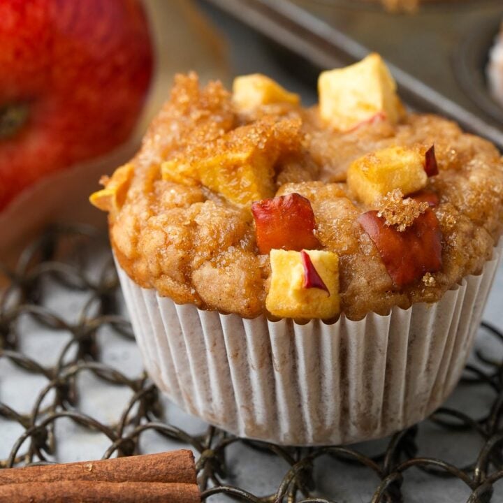 A close-up of a Vegan Apple Muffin topped with apple pieces and cinnamon, with an apple and a cinnamon stick in the background on a metal cooling rack.