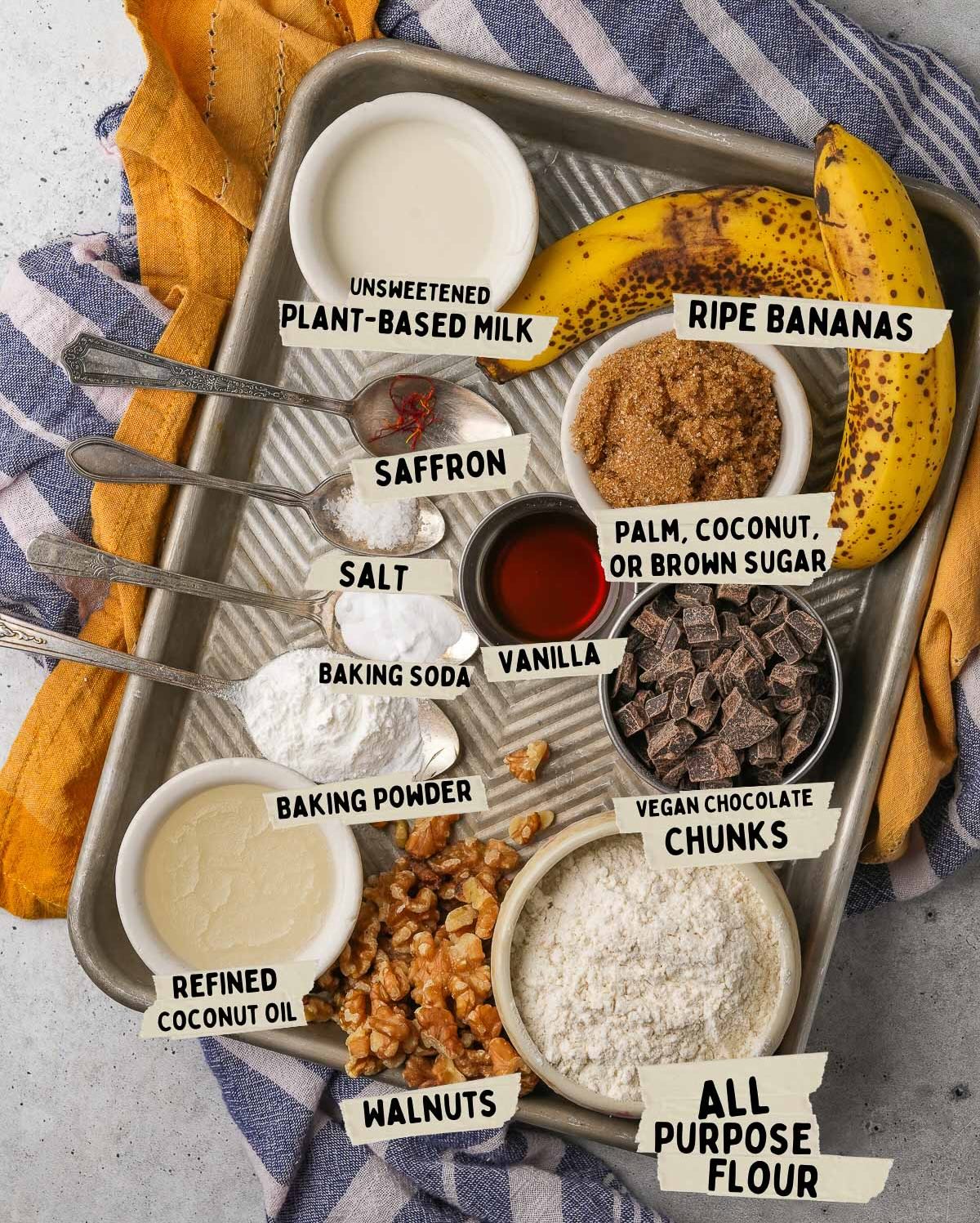 A baking tray contains labeled ingredients for vegan banana muffins, including plant-based milk, ripe bananas, sugar, vanilla, chocolate chunks, flour, walnuts, coconut oil, baking powder, baking soda, saffron, and salt.