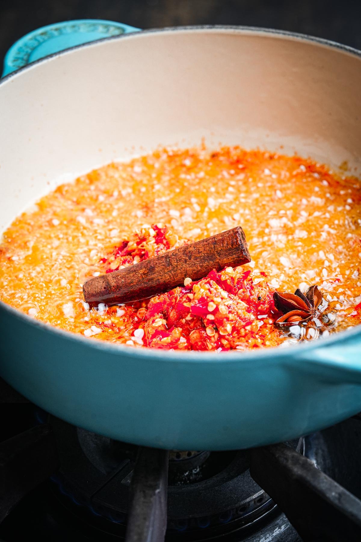 A light blue pot on a stove containing chopped red chili peppers, a cinnamon stick, and star anise, all being cooked together.
