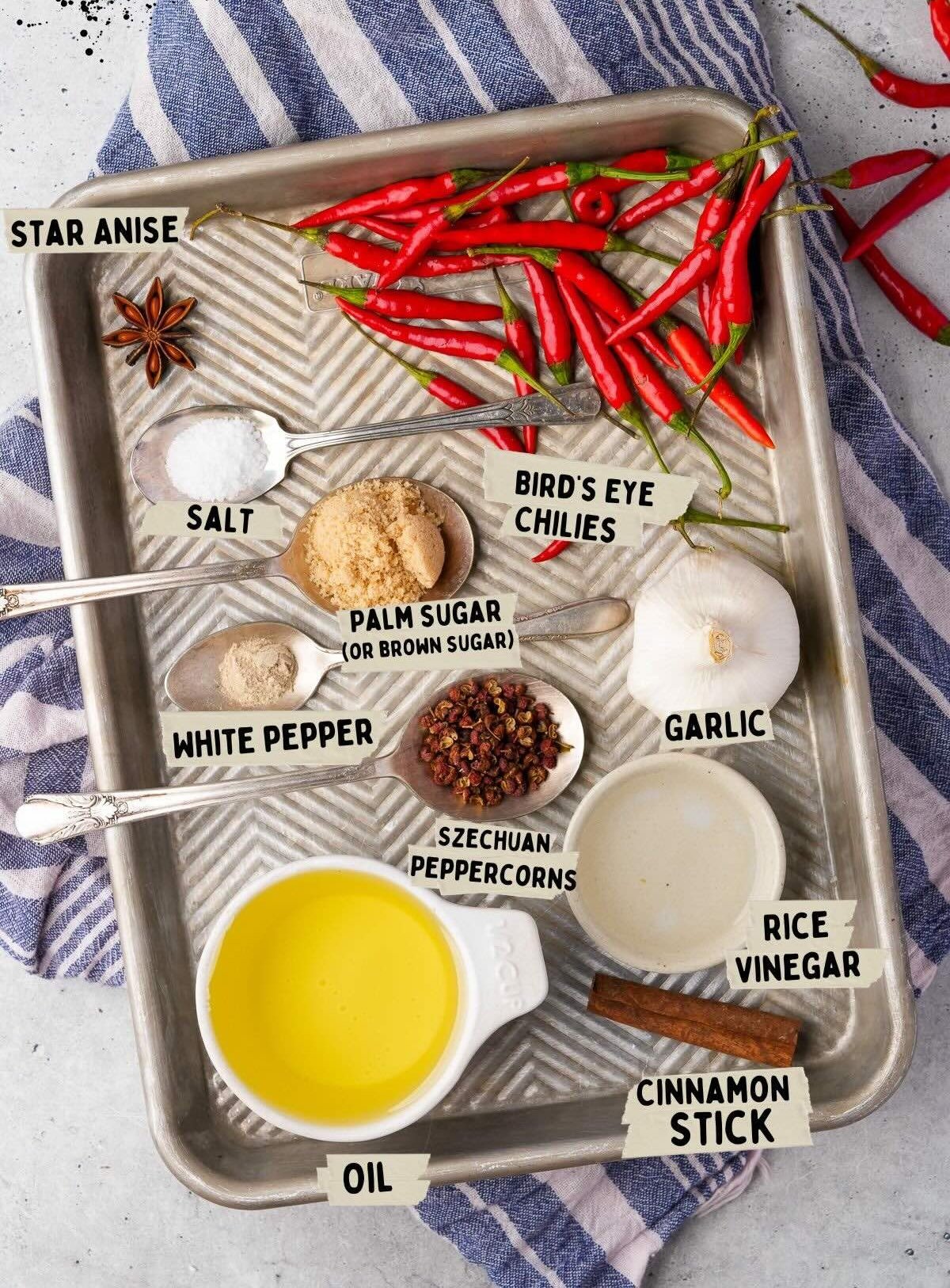 A tray with bird's eye chilies, garlic, cinnamon stick, star anise, white pepper, salt, oil, rice vinegar, Szechuan peppercorns, a brown sugar alternative, and a jar of chili garlic sauce—all labeled accordingly.