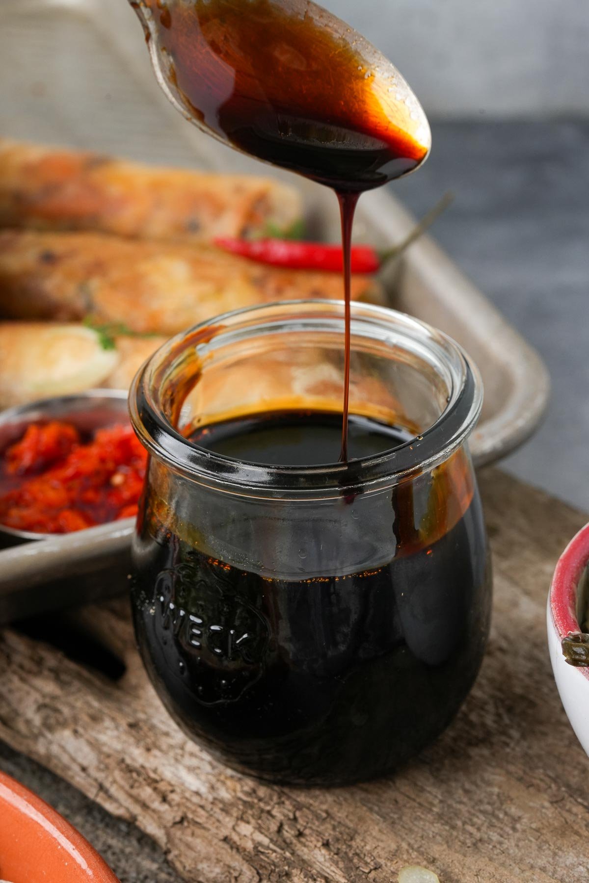 A spoon drizzles dark kecap manis into a glass jar. Various dishes, including spring rolls, are visible in the background.