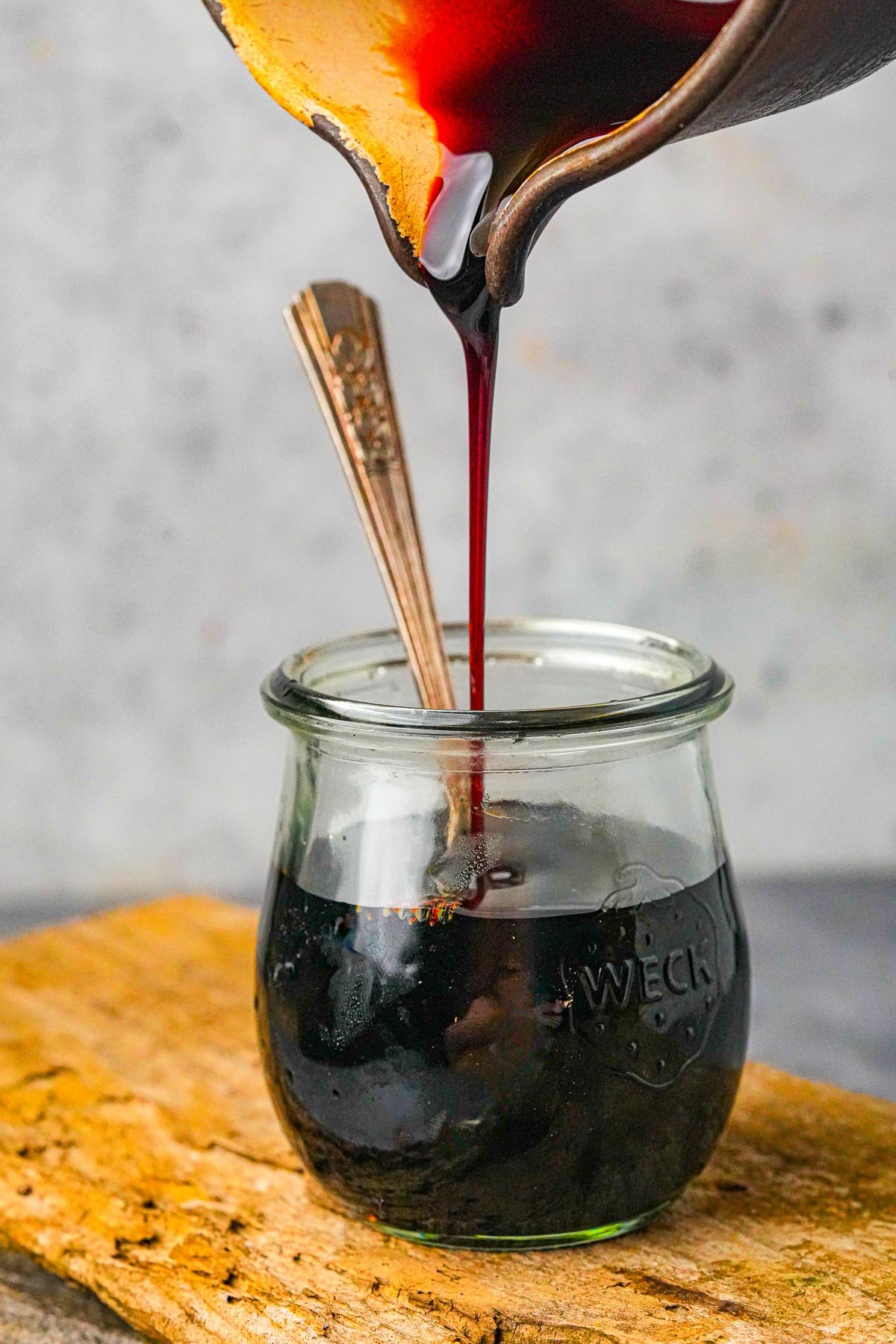 Pouring kecap manis from a pot into a small glass jar with a spoon inside, placed on a wooden surface.