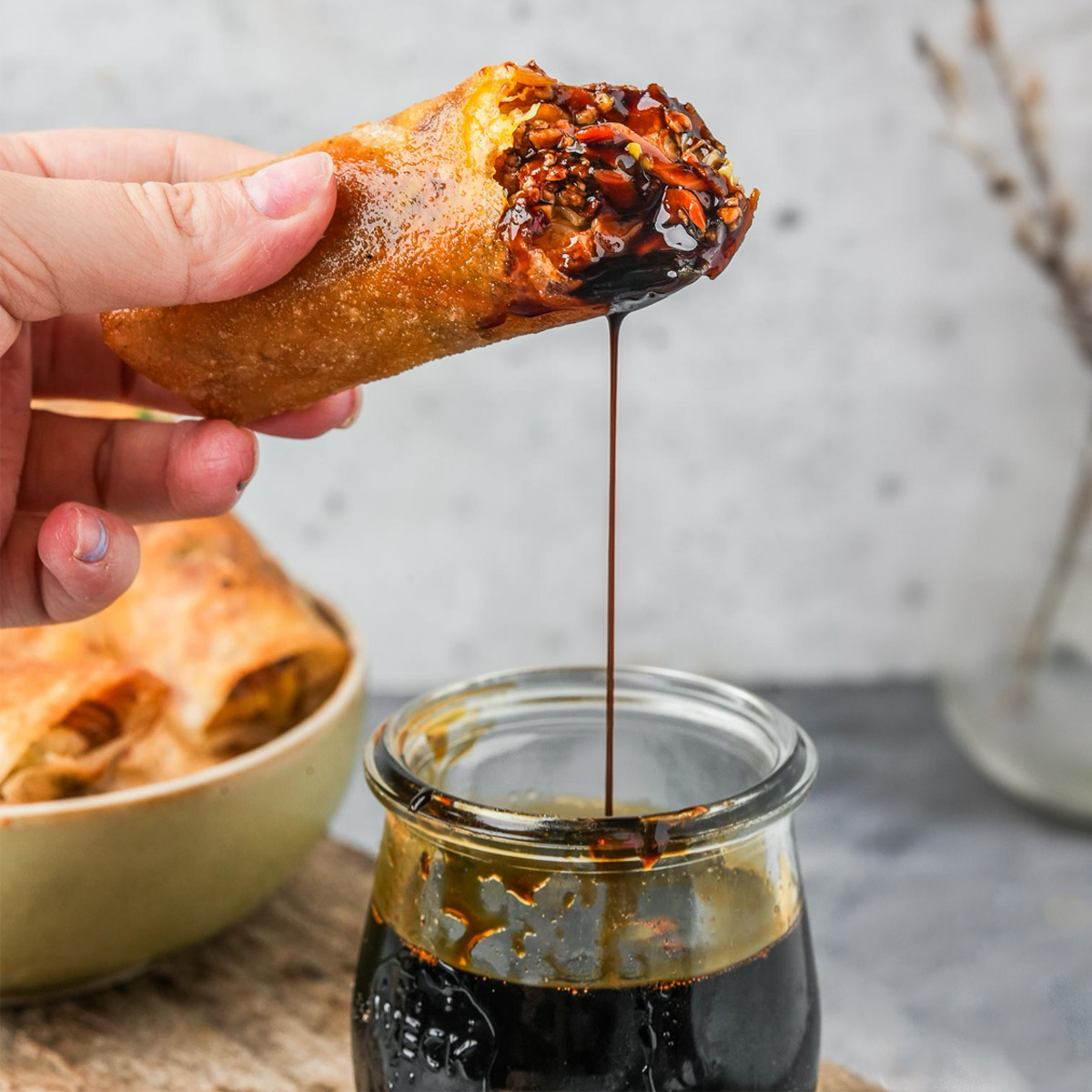 Hand dipping a spring roll into a glass jar filled with thick, dark kecap manis. A plate with more spring rolls is visible in the background.