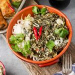 A bowl of white rice and Filipino Laing topped with red chili peppers, and cilantro, placed on a wooden surface with a fork and rolls in the background.