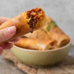 Close-up of a hand holding a Lumpiang Shanghai roll filled with vegetables and soy sauce, with more spring rolls in a bowl in the background.