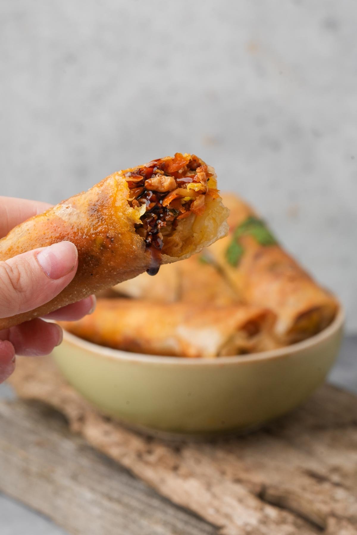 A hand holds a half-eaten Lumpiang Shanghai roll filled with vegetables above a bowl containing more spring rolls, placed on a wooden surface.