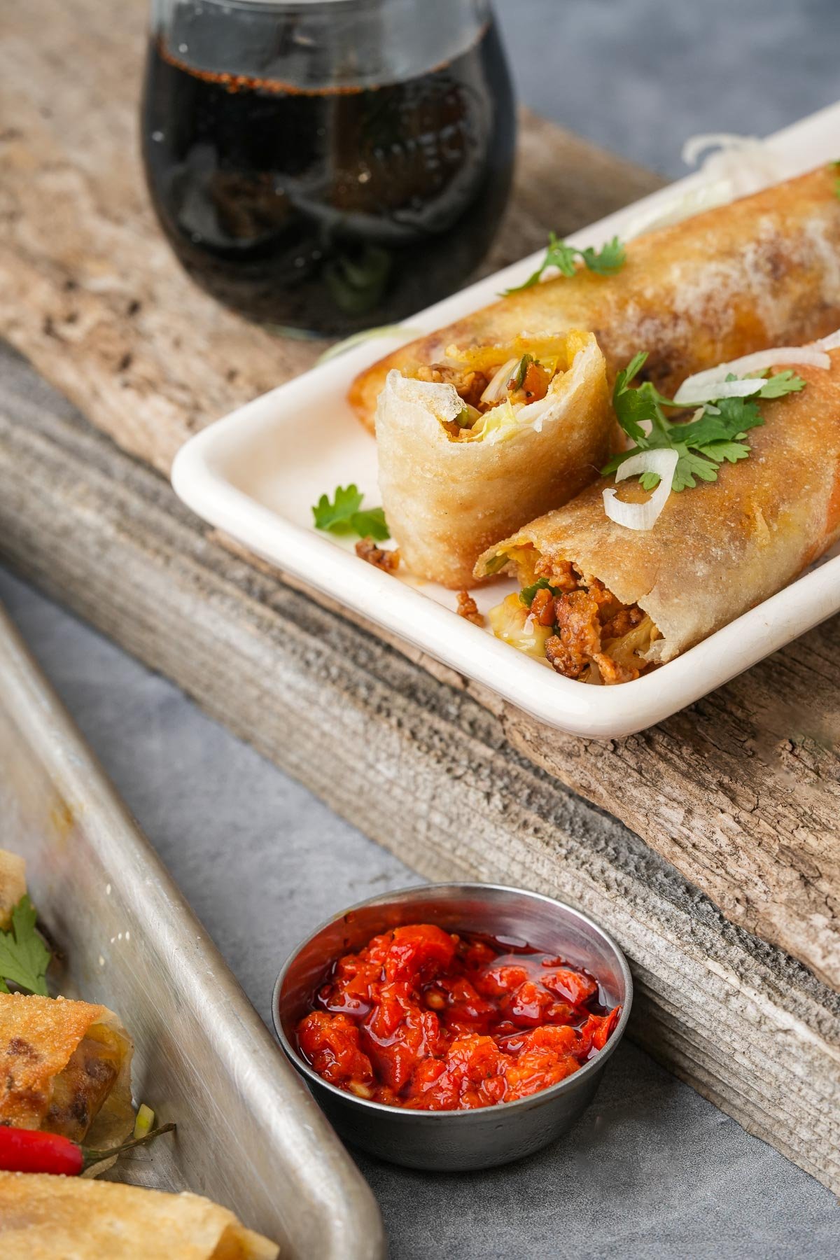 Two Lumpiang Shanghai rolls on a white plate, garnished with coriander, beside a small bowl of chili sauce and a glass with kecap manis in it.