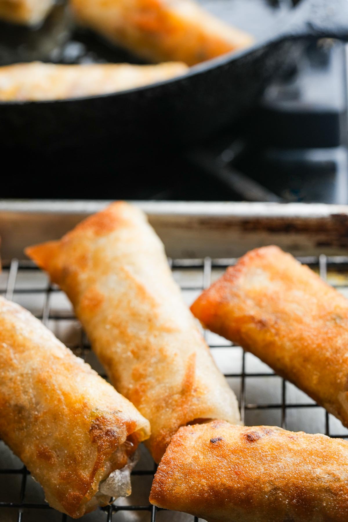 Four golden-brown Lumpiang Shanghai rolls rest on a cooling rack with a skillet cooking more rolls in the background.