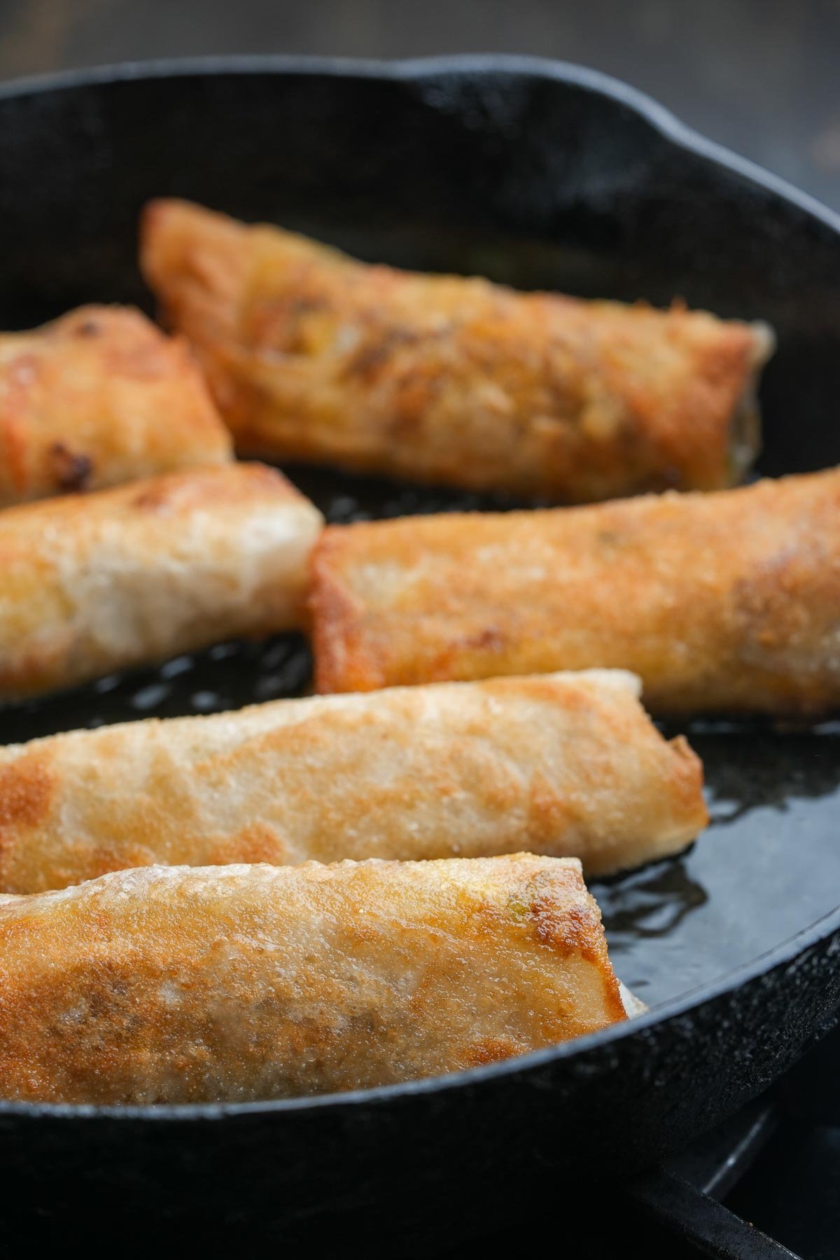 Six golden-brown Lumpiang Shanghai rolls frying in a black cast iron skillet.