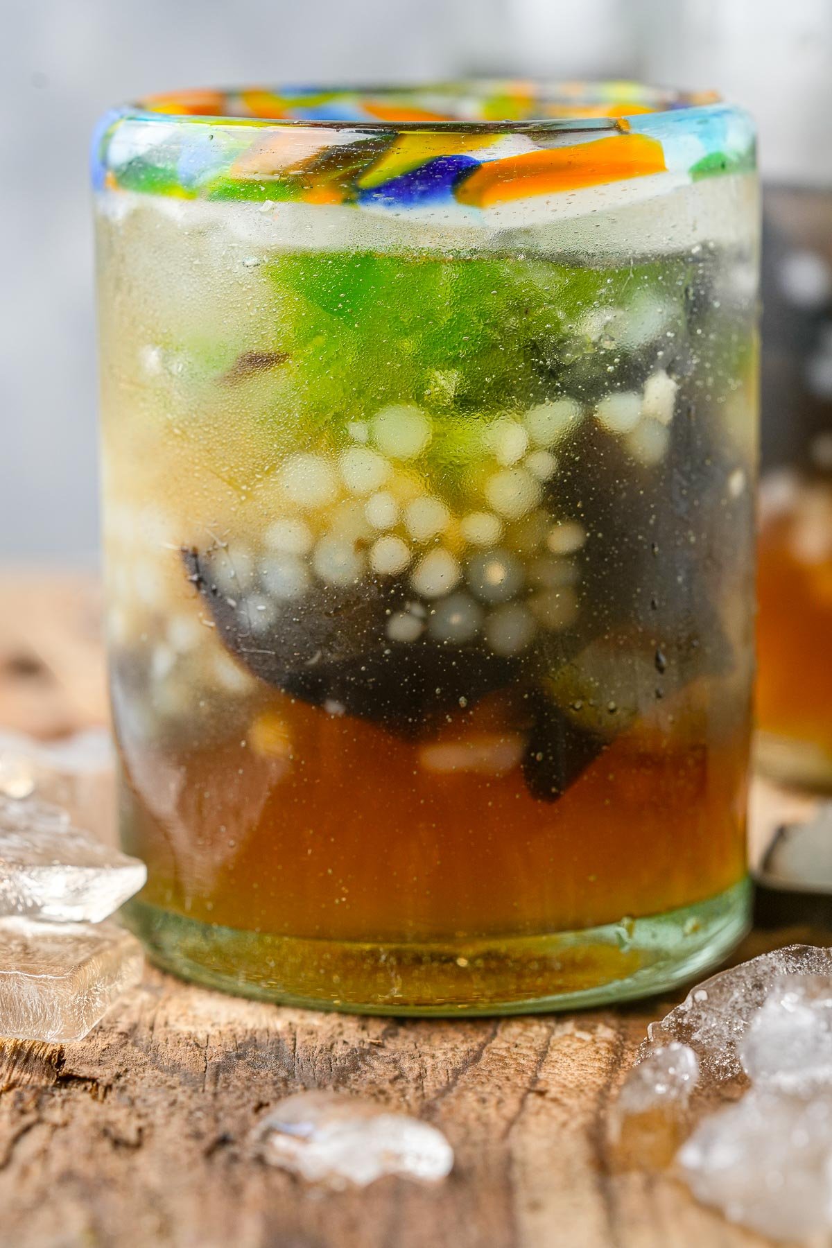 A close-up of Sagot Gulaman with bubbles, condensation and various jellies inside a glass, placed on a wooden surface with ice cubes scattered around.