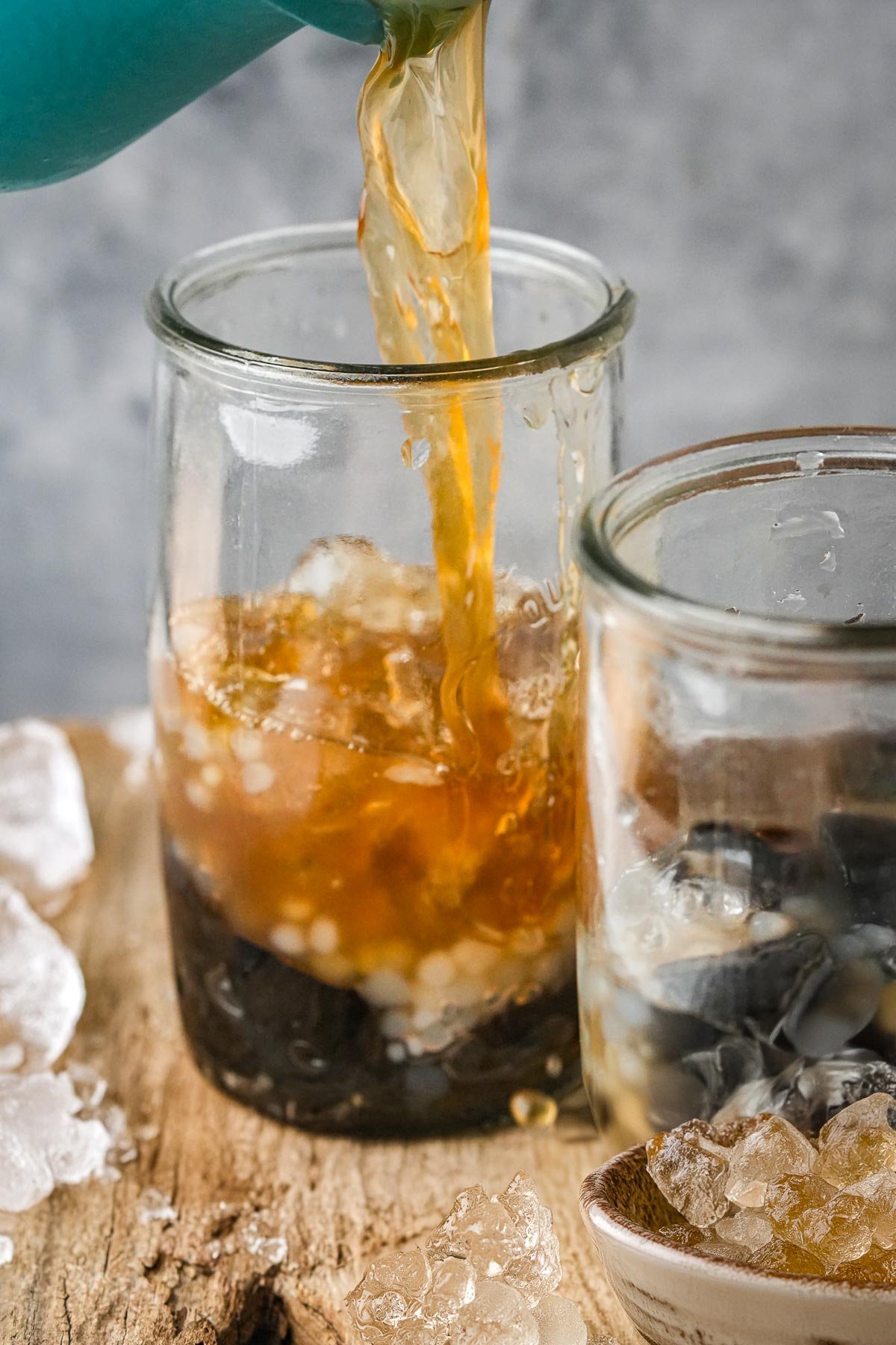 Arnibal syrup is being poured into a glass containing dark jelly pieces, next to another glass with similar contents on a wooden surface, with ice cubes scattered around.