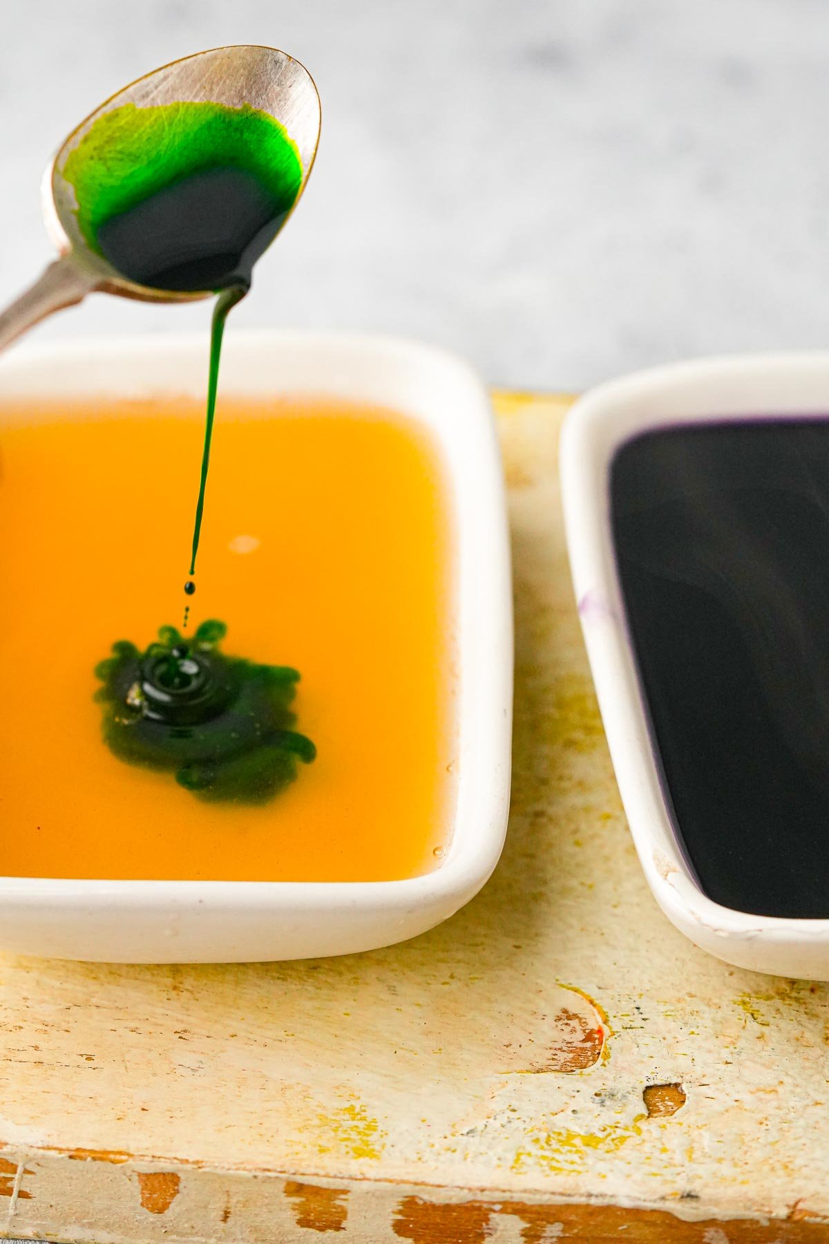 A spoon is dripping green pandan extract into a small rectangular white dish containing liquid jelly that still hasnt set up yet.