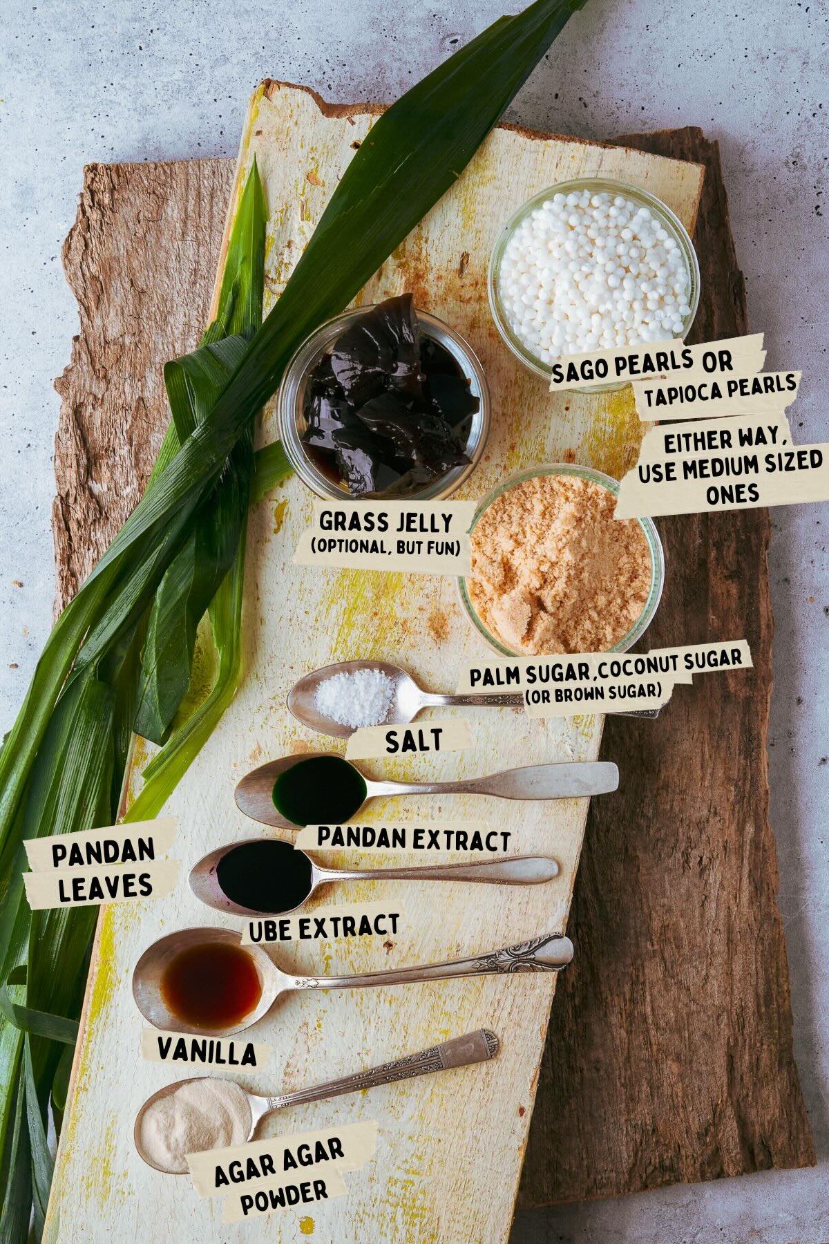 Ingredients for a Sagot Gulaman recipe laid out on a wooden board, including pandan leaves, grass jelly, sago pearls, palm sugar, salt, pandan extract, ube extract, vanilla, and agar-agar powder.