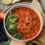 A bowl of red salsa rojo garnished with cilantro, surrounded by tortilla chips, a slice of tomato, lime wedge, and dried pepper on a wooden surface.