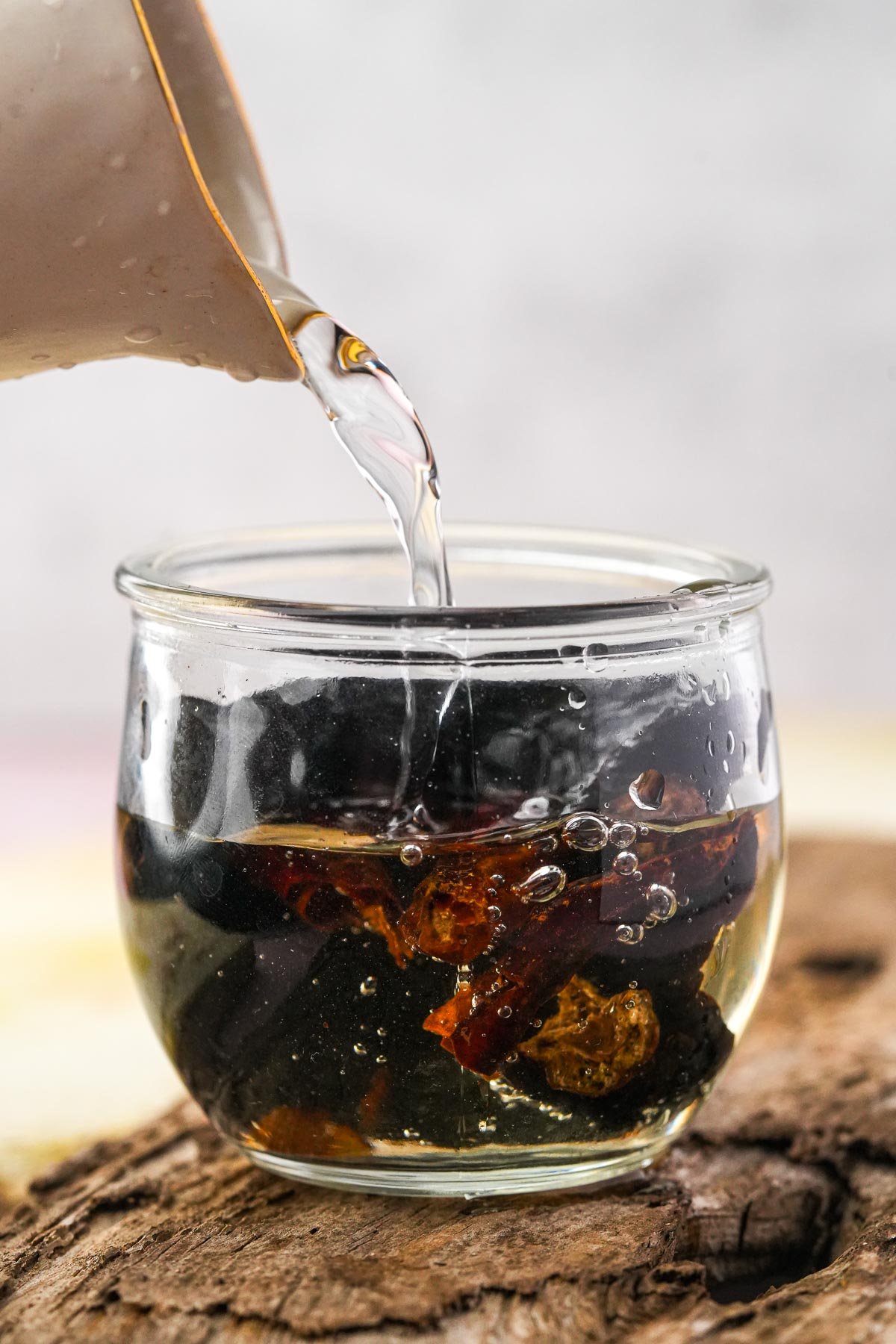 Water is being poured from a beige pitcher into a transparent glass jar containing dried chilies. The jar is placed on a wooden surface.