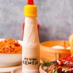 Bottle of vegan Kewpie mayonnaise with a yellow cap placed in a white bowl, surrounded by dishes of food including noodles and a serving of seitan with vegetables, set against a grey background.