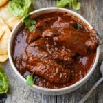 A bowl filled with red Adobo Sauce with chipotle peppers, garnished with cilantro, placed on a wooden surface near some tortilla chips and lettuce leaves.