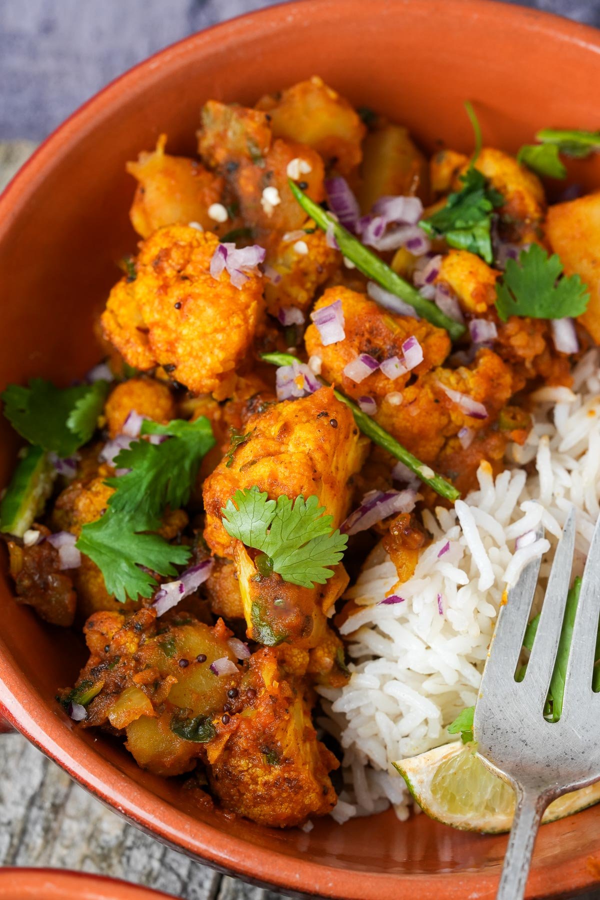 A close-up view of a bowl containing rice and Aloo Gobi garnished with herbs, served with a fork and a wedge of lemon.