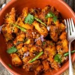 A bowl of Aloo Gobi garnished with cilantro, green chilies, chopped onions, and sev, with a fork on the side.