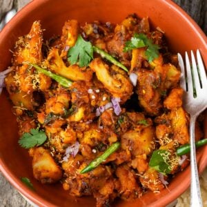A bowl of Aloo Gobi garnished with cilantro, green chilies, chopped onions, and sev, with a fork on the side.