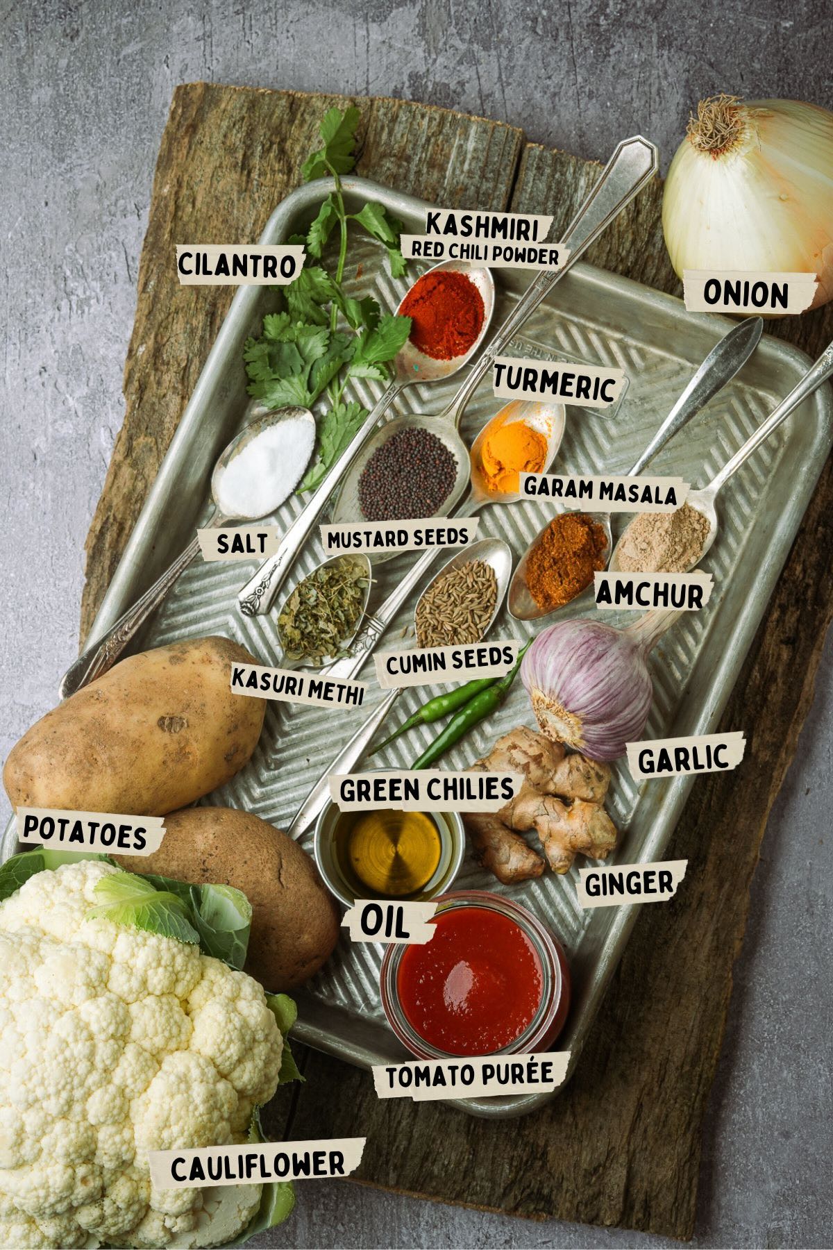 A tray with various spices and ingredients for making Aloo Gobi including cilantro, kashmiri chili powder, onion, turmeric, garam masala, salt, mustard seeds, amchur, kasuri methi, cumin seeds, garlic, green chilies, ginger, potatoes, oil, tomato puree, and cauliflower.