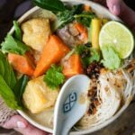 A bowl of cha ri chay with vegetables, tofu, herbs, lime, and a ceramic spoon, held by a young woman.