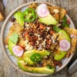 A bowl of chilaquiles topped with sliced avocado, radishes, jalapeños, crumbled vegan cheese, cilantro, and a spicy red sauce on a wooden surface.