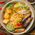 A bowl of Curry Laksa with various toppings such as tofu puffs, lime slices, bean sprouts, cilantro, chilies, and fried eggplant.