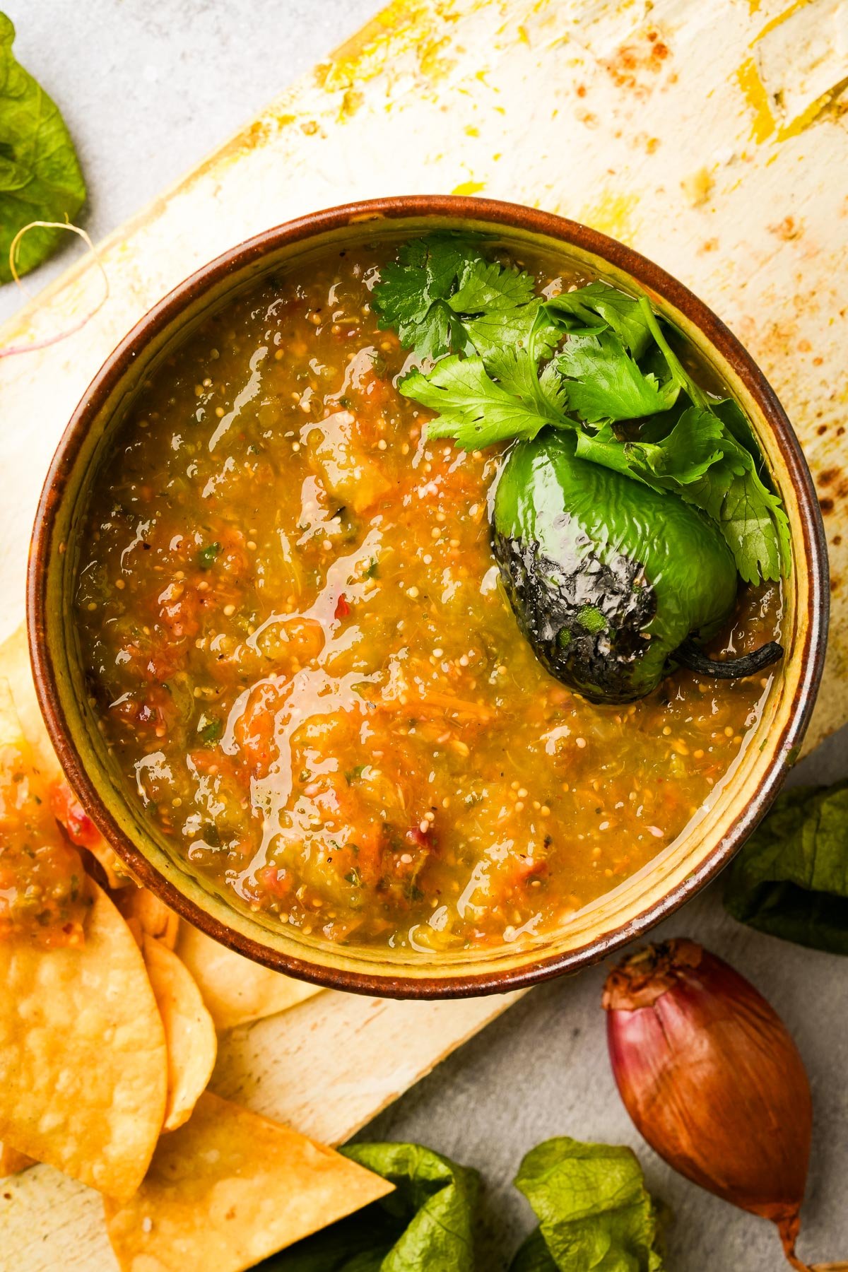 A bowl of salsa verde garnished with cilantro and a roasted jalapeño sits on a wooden board. Nearby are tortilla chips, an onion, and green leaves.