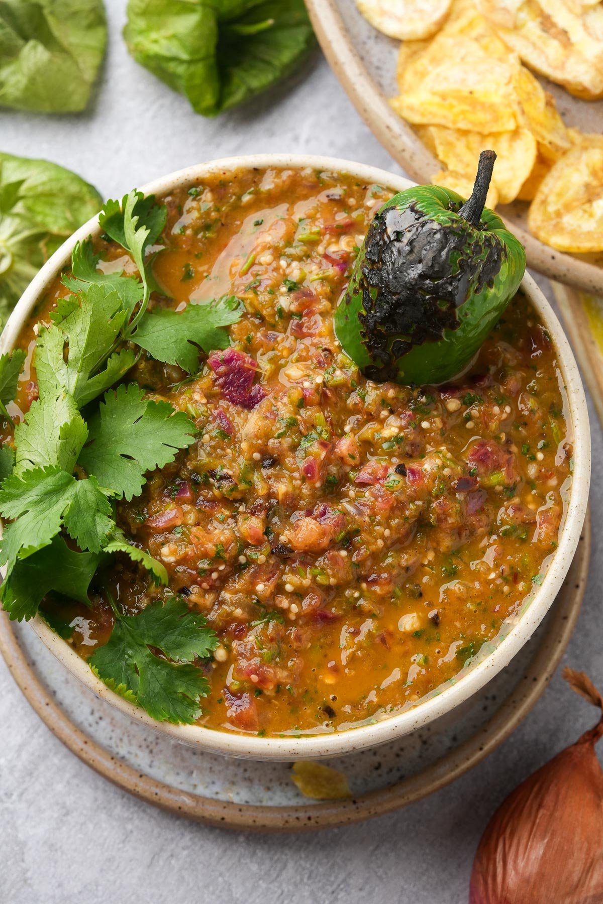 A bowl of salsa verde garnished with a charred jalapeño and fresh cilantro, placed on a plate next to sliced plantain chips.