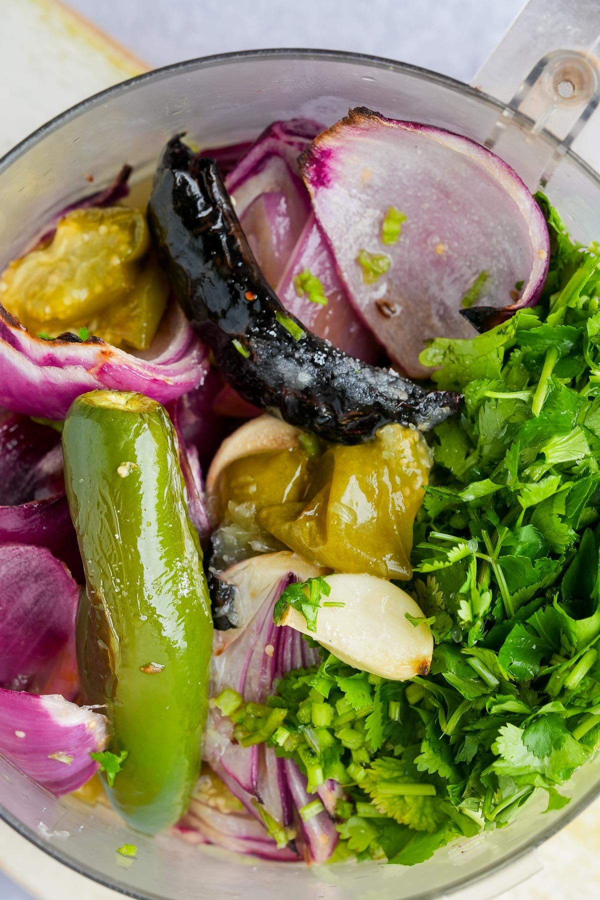 Ingredients for making salsa verde in a food processor, including roasted onions, green chili peppers, garlic, and fresh cilantro, ready for blending.
