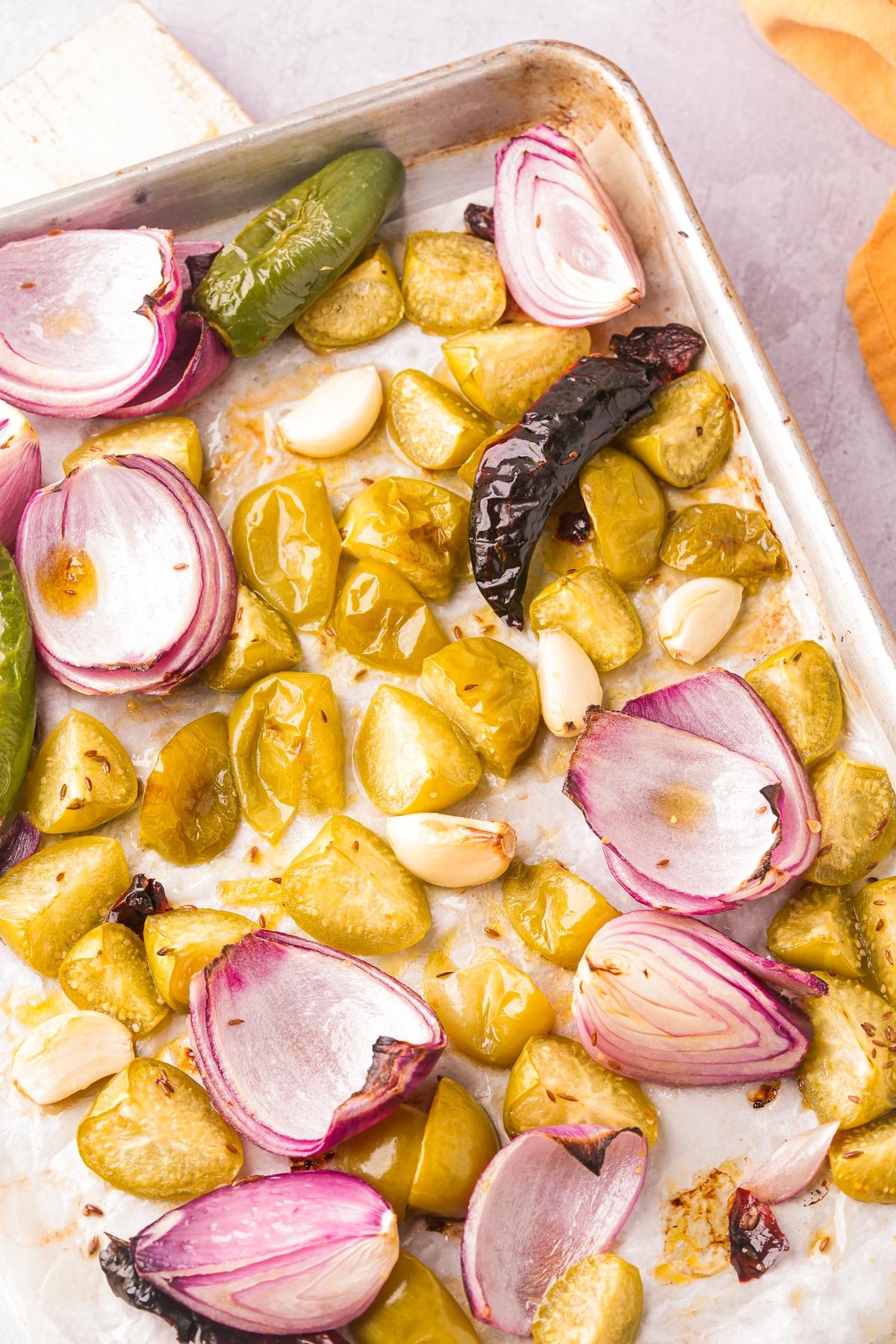 A baking sheet containing roasted vegetables, including halved onions, yellow tomatoes, garlic cloves, green chili peppers, and dried red chilies.