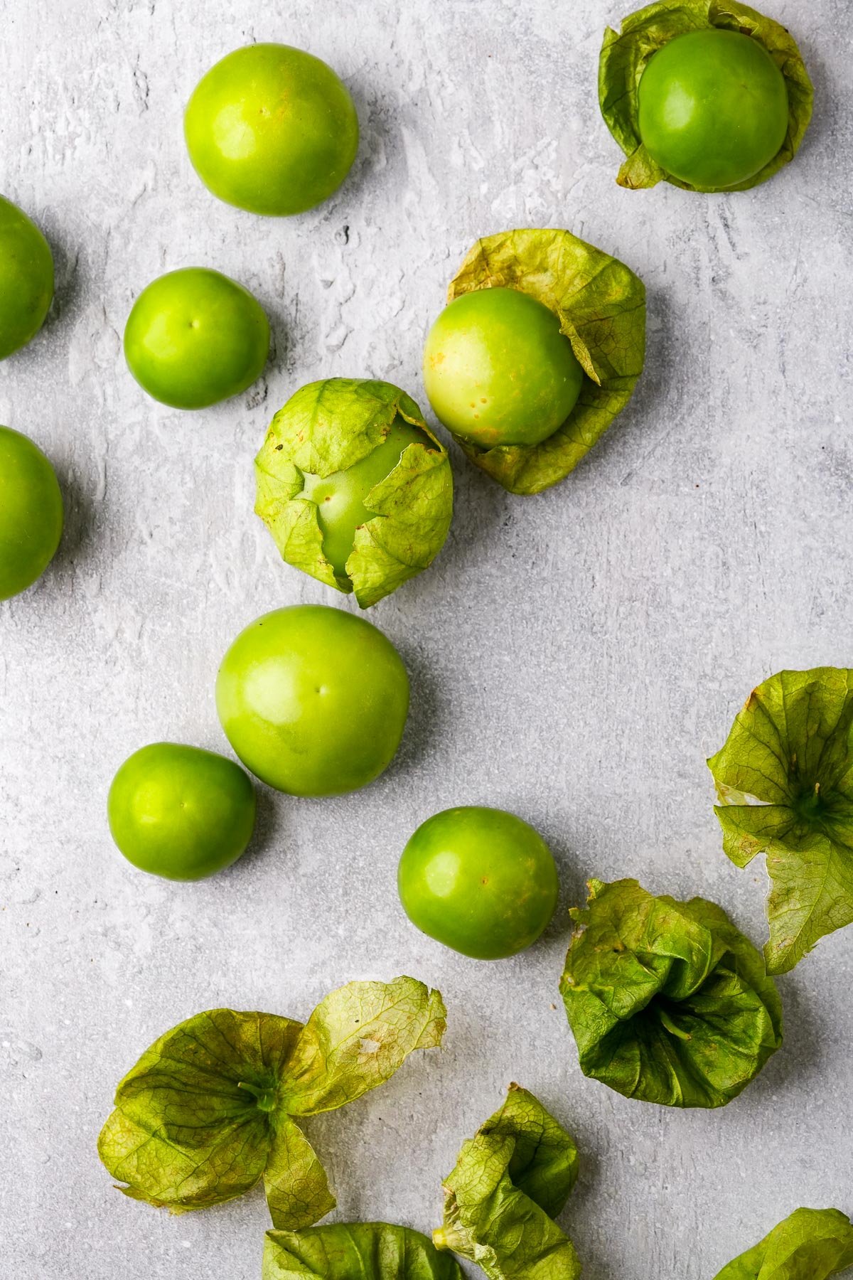 Several fresh green tomatillos, some still in their husks, are scattered on a light gray textured surface.