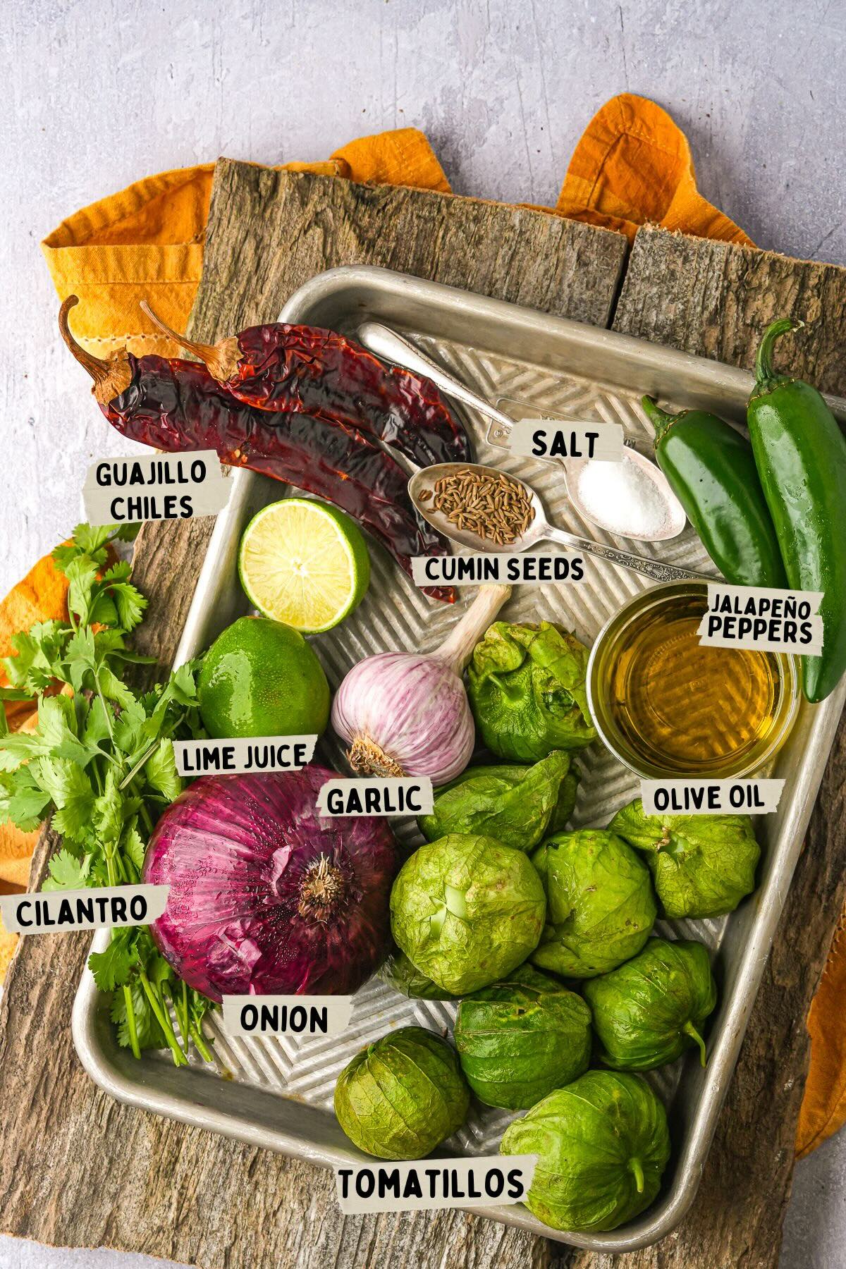 A tray containing ingredients for making salsa verde, including guajillo chiles, jalapeño peppers, tomatillos, onion, garlic, cilantro, lime juice, cumin seeds, salt, and olive oil.