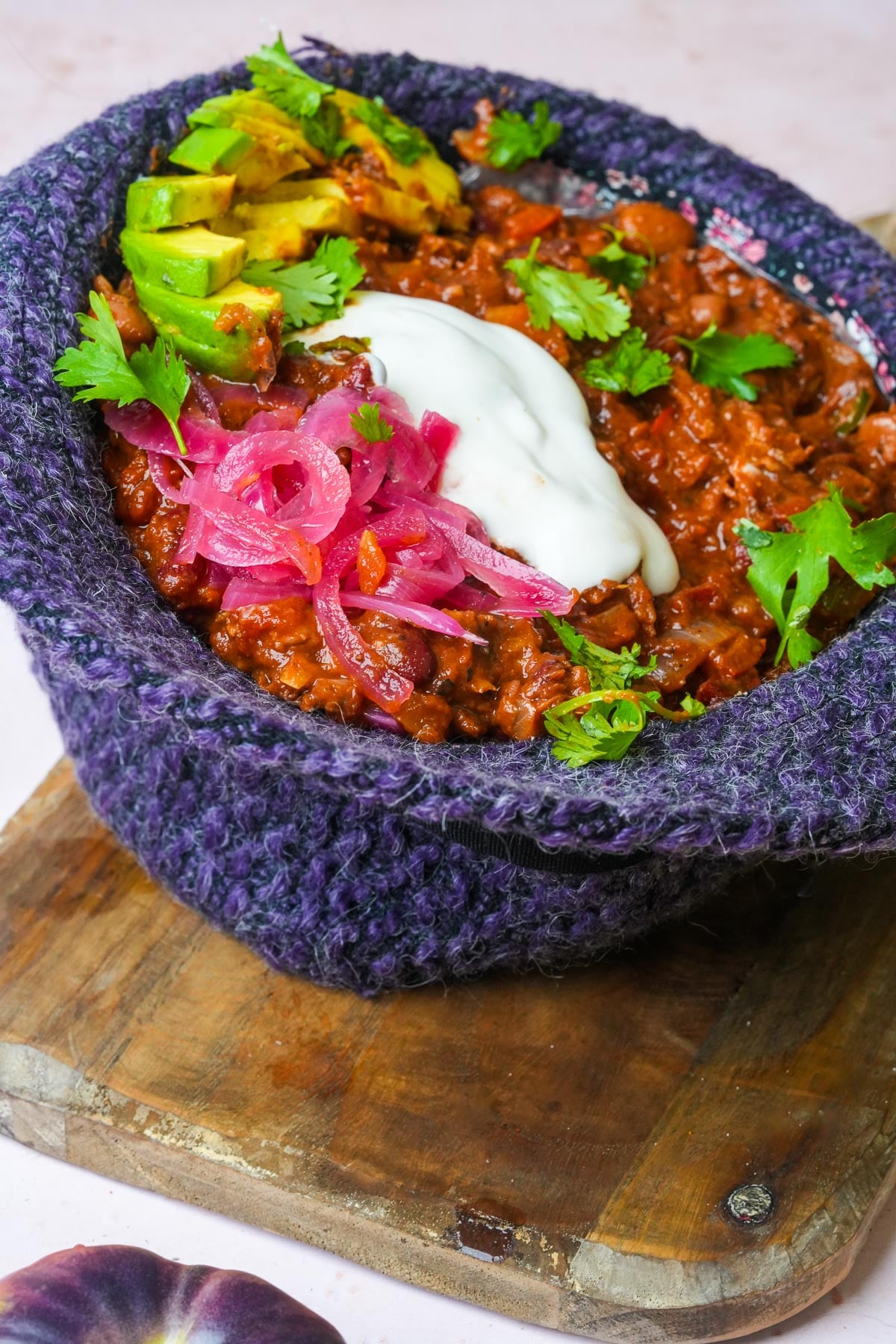 A hat full of vegan chili topped with sliced avocado, pickled onions, cilantro, and sour cream, placed on a wooden board.