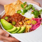 A person holds a brimmed hat filled with vegan chili topped with avocado slices, shredded cheese, sour cream, pickled onions, jalapeño slices, cilantro, and tortilla chips.