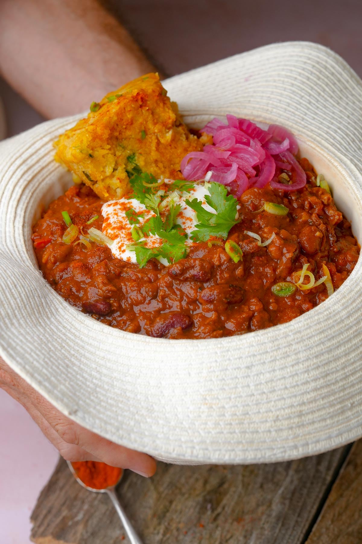 A person holding a hat of vegan chili topped with vegan sour cream, cilantro, pickled onions, and cornbread, served in a white hat.